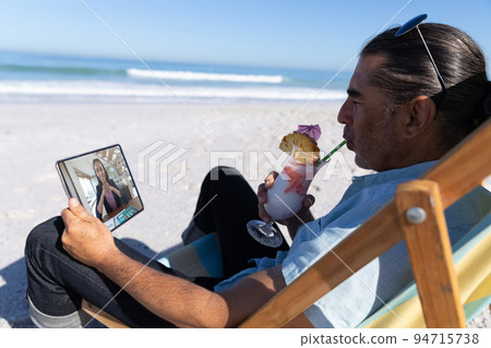 Caucasian man relaxing on beach with drink having video call using tablet 94715738