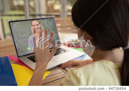Caucasian schoolgirl wearing face mask and waving on laptop video call with female teacher 94716934