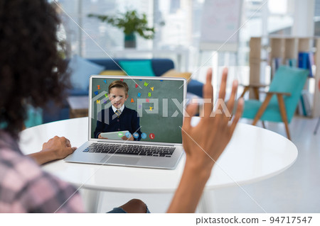Mixed race female teacher using laptop and waving on video call with schoolboy 94717547