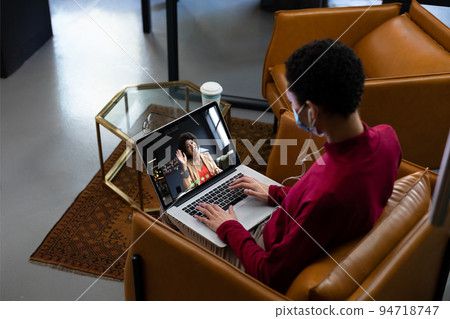 African american woman wearing face mask having video call with female colleague displayed on laptop 94718747