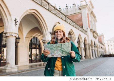 Attractive young female tourist is exploring new city. Redhead girl in hat holding a paper map on Market Square in Krakow. Traveling Europe in autumn. High quality photo 94726305