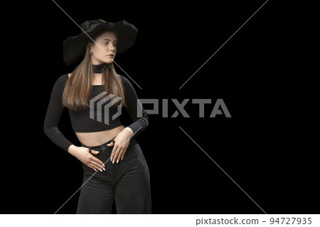 Young girl in short top, jeans and wide-brimmed hat poses in the studio on black background. Copy space. 94727935