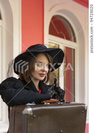 Portrait of beautiful aristocratic woman in black wide-brimmed hat and lace gloves with retro suitcase. Female traveler Portrait of beautiful aristocratic woman in black wide-brimmed hat and lace gloves with retro suitcase. Female traveler 94728020