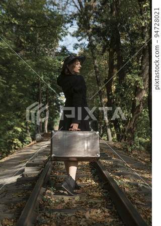 Woman in black retro clothes walks with a suitcase along the rails through the autumn forest. View from the back Woman in black retro clothes walks with a suitcase along the rails through the autumn forest. View from the back 94728021