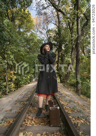 Stylish woman in black coat and broad-brim hat stands near vintage suitcase on rails in autumn wood. Vertical frame Stylish woman in black coat and broad-brim hat stands near vintage suitcase on rails in autumn wood. Vertical frame 94728026