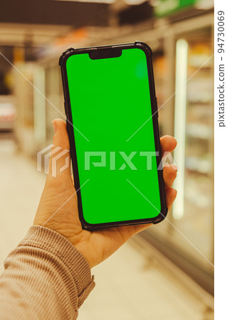 Mobile phone Chroma key on defocused frozen food fridge background. Close up of woman hand holds smart phone with green screen at shopping mall store. Buying food at supermarket, grocery shop center Mobile phone Chroma key on defocused frozen food fridge background. Close up of woman hand holds smart phone with green screen at shopping mall store. Buying food at supermarket, grocery shop center 94730069