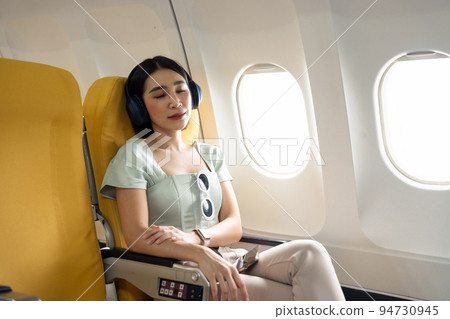 Tired asian lady napping with headphones on seat while traveling by airplane. Commercial transportation by planes. 94730945