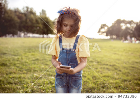 little girl 5 years old in yellow blouse and denim overalls smiles into smartphone on sunny day in park 94732386