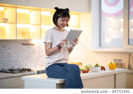 Young woman with a gadget sitting in the kitchen Young woman with a gadget sitting in the kitchen 94732492