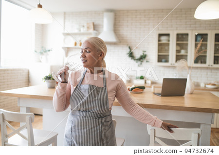 Female in the apron sipping a beverage in the kitchen 94732674