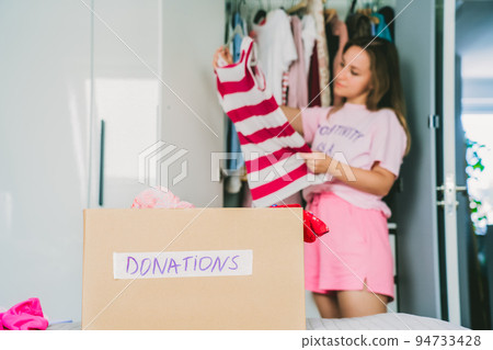 A cardboard box of selected clothes for donating to a Charity shop standing on the bed. Decluttering, Sorting clothes, and Cleaning Up. Reuse, second-hand. Conscious consumer, sustainability. 94733428