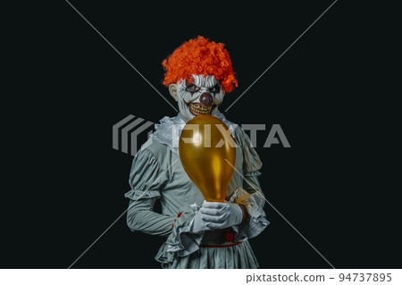 evil clown holds a golden balloon in his hands 94737895