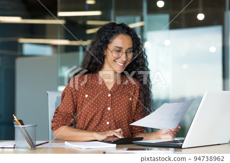 Young successful businesswoman behind paperwork, Hispanic woman working with documents and contracts inside modern office building, female worker using laptop, wearing glasses and curly hair smiling. Young successful businesswoman behind paperwork, Hispanic woman working with documents and contracts inside modern office building, female worker using laptop, wearing glasses and curly hair smiling. 94738962