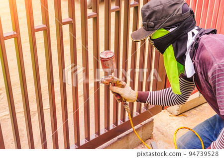 Male workers are using a paint sprayer at a steel door. Workers spray paint must be protected from inhalation by using cloth to cover the nose. Male workers are using a paint sprayer at a steel door. Workers spray paint must be protected from inhalation by using cloth to cover the nose. 94739528