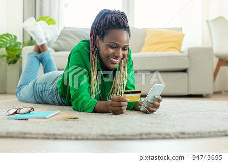 Excited black woman laying on floor carpet with cell phone and plastic credit card, ordering food or shopping online 94743695