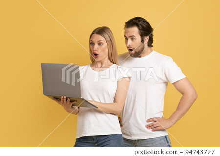 Shocked young caucasian couple in white t-shirts with open mouths look at laptop 94743727