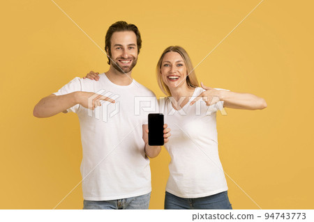 Glad happy young caucasian couple in white t-shirts point fingers at smartphone with blank screen and recommend app Glad happy young caucasian couple in white t-shirts point fingers at smartphone with blank screen and recommend app 94743773