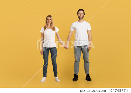Smiling young caucasian female and male in white t-shirts hold hands, enjoy spare time, look at camera 94743774