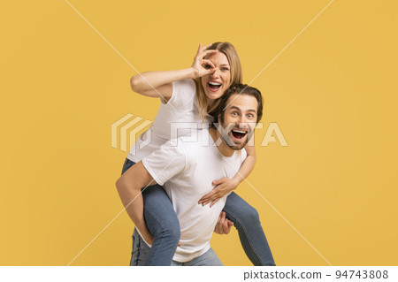 Happy young caucasian guy in white t-shirt holding lady on back, have fun, isolated on yellow background Happy young caucasian guy in white t-shirt holding lady on back, have fun, isolated on yellow background 94743808