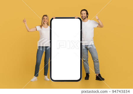 Satisfied young caucasian couple in white t-shirts show thumbs up near huge smartphone with blank screen Satisfied young caucasian couple in white t-shirts show thumbs up near huge smartphone with blank screen 94743834