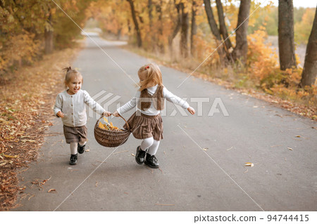 Amusing funny laughing girls are running along the road with a basket full of yellow dry fallen leaves. Children collect autumn foliage Amusing funny laughing girls are running along the road with a basket full of yellow dry fallen leaves. Children collect autumn foliage 94744415