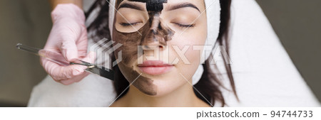 cosmetologist applying black mask on pretty woman face wearing black gloves, gorgeous woman in spa having facial procedures 94744733