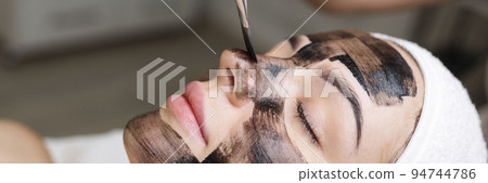 cosmetologist applying black mask on pretty woman face wearing black gloves, gorgeous woman in spa having facial procedures 94744786