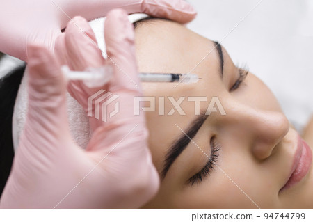 Close up of hands of young cosmetologist injecting botox in female face. She is standing and smiling. The woman is closed her eyes with relaxation 94744799