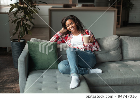 Cheerful millennial african american lady with cup of favorite drink, enjoy free time and comfort, sit on sofa Cheerful millennial african american lady with cup of favorite drink, enjoy free time and comfort, sit on sofa 94747010