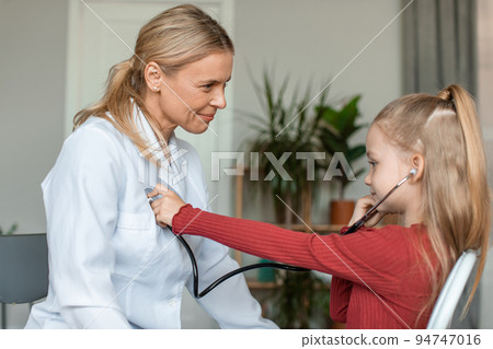 Pretty little girl visiting pediatrician and playing with female doctor, listening doc's heart beating with stethoscope 94747016