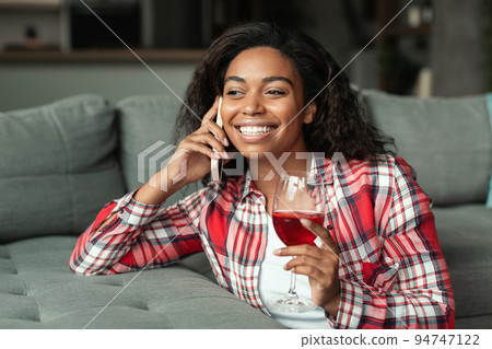 Happy millennial african american woman with glass of wine talking by smartphone in living room interior Happy millennial african american woman with glass of wine talking by smartphone in living room interior 94747122