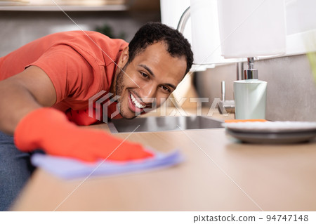 Smiling millennial black male in rubber gloves wipes dust from wooden table in kitchen interior, free space 94747148