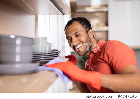Glad millennial black man in rubber gloves wipes dust from shelf with dishes enjoy purely in white kitchen Glad millennial black man in rubber gloves wipes dust from shelf with dishes enjoy purely in white kitchen 94747149