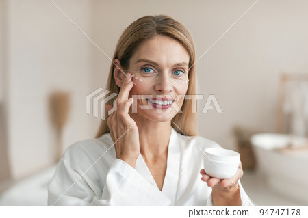 Beautiful middle aged woman in silky bathrobe using moisturizing eye cream, enjoying face care routine Beautiful middle aged woman in silky bathrobe using moisturizing eye cream, enjoying face care routine 94747178