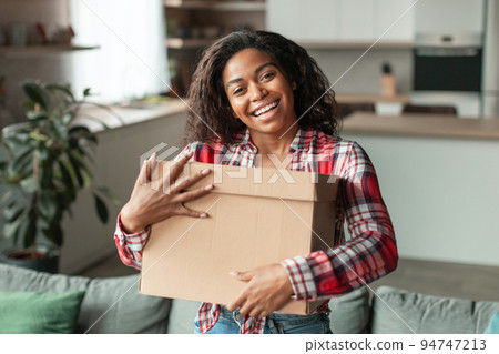 Satisfied pretty millennial african american lady hugs cardboard box with purchases in living room Satisfied pretty millennial african american lady hugs cardboard box with purchases in living room 94747213