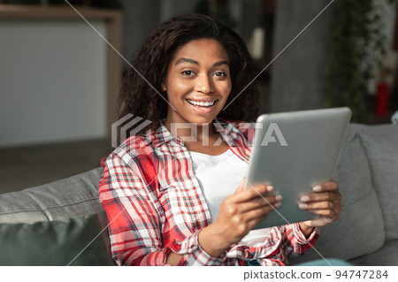 Surprised happy millennial african american woman typing on tablet on sofa in living room interior 94747284