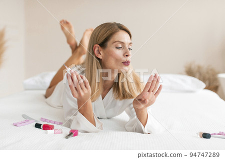 Gorgeous middle aged woman doing manicure, applying nail polish and blowing on nails while lying on bed at home Gorgeous middle aged woman doing manicure, applying nail polish and blowing on nails while lying on bed at home 94747289