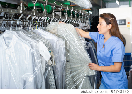Adult female worker of laundry inspecting clothing after cleaning on racks 94748180