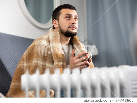 Young man wrapped in plaid sitting on sofa near radiator with cup 94748603