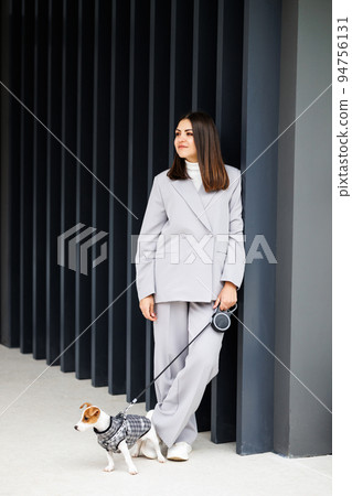 Woman in casual jacket standing with her favorite fluffy trained purebred Jack Russell Terrier dog dressed in suit for dogs near wall in outdoor in autumn day. Woman in casual jacket standing with her favorite fluffy trained purebred Jack Russell Terrier dog dressed in suit for dogs near wall in outdoor in autumn day. 94756131