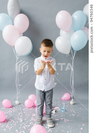 Happy birthday, child. Photo of adorable cute baby little boy blowing on confetti on the background of balloons Happy birthday, child. Photo of adorable cute baby little boy blowing on confetti on the background of balloons 94760346