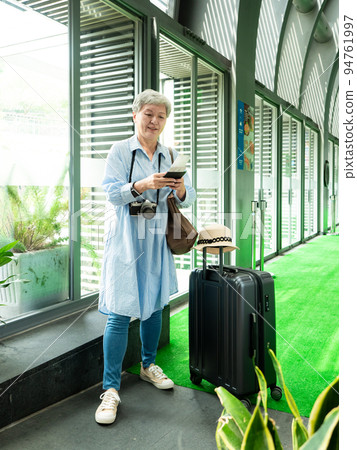 60多歲的亞洲熟年婦女微笑著站在身旁有手提箱行李袋和使用智能手機,適用於covid19疫情後旅行的概 60多歲的亞洲熟年婦女微笑著站在身旁有手提箱行李袋和使用智能手機,適用於covid19疫情後旅行的概 94761997