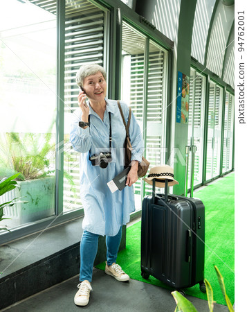 60多歲的亞洲熟年婦女微笑著站在身旁有手提箱行李袋和使用智能手機,適用於covid19疫情後旅行的概 94762001