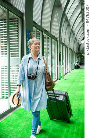 60多歲的亞洲熟年婦女微笑著站著,身旁有手提箱行李袋和手持草帽,適用於covid19疫情後旅行的概念 94762007