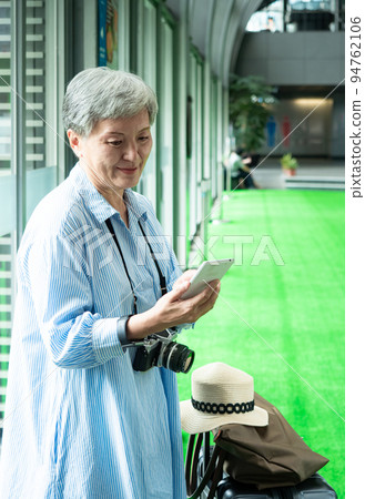 60多歲的亞洲熟年婦女微笑著站在身旁有手提箱行李袋和使用智能手機,適用於covid19疫情後旅行的概 94762106