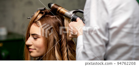 The female hairdresser is curling hair for a brown-haired young caucasian woman in a beauty salon 94762208