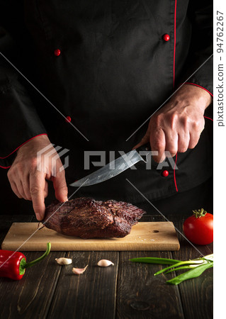 Professional chef cuts the baked beef steak with a knife on a wooden board. The concept of the cooking process food for hotel Professional chef cuts the baked beef steak with a knife on a wooden board. The concept of the cooking process food for hotel 94762267