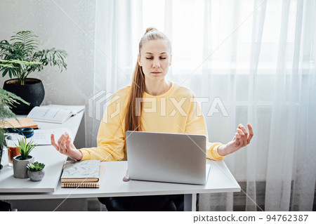 Young woman with close eyes near laptop at home office practicing meditation at table. Freelancer woman meditating near laptop. Taking break time during work. Healing from paperwork Young woman with close eyes near laptop at home office practicing meditation at table. Freelancer woman meditating near laptop. Taking break time during work. Healing from paperwork 94762387