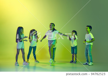 Dance class. Joyful children, kids in t-shirts and jeans dancing in action isolated on green background in yellow neon light. Dance class. Joyful children, kids in t-shirts and jeans dancing in action isolated on green background in yellow neon light. 94762523