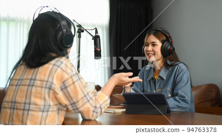 Smiling female radio host discussing various topics with her guest while streaming live audio podcast from home studio Smiling female radio host discussing various topics with her guest while streaming live audio podcast from home studio 94763378